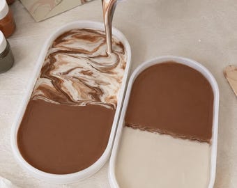 May include: Two oval white silicone molds being filled with liquid casting material. One mold contains a swirling mixture of brown and white liquid, while the other holds distinct halves of solid brown and white material. A Marble Tray Kit box is visible.