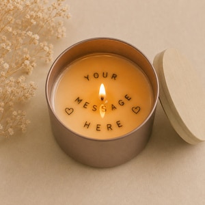 May include: A lit candle in a rose gold tin with the words "YOUR MESSAGE HERE" and heart symbols. The candle has a wooden lid and is surrounded by dried flowers. The candle is on a light beige surface.
