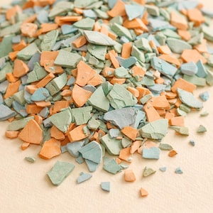 Può includere: Un mucchio di pezzi rotti, di forma irregolare, nei toni dell'arancione, del verde e del blu. I frammenti sono sparsi su una superficie beige chiaro, creando un'esposizione strutturata e colorata. I pezzi sembrano essere fatti di un materiale solido.