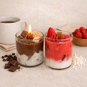 Può includere: Due candele a tema dessert in barattoli di vetro trasparente. Una candela ha una copertura di cioccolato e waffle, l'altra ha una copertura di lamponi. In ognuna è visibile uno stoppino di candela acceso. Una tazza di caffè, chicchi di caffè e una ciotola di lamponi sono anch'essi nell'immagine.