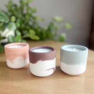 Può includere: Tre candele artigianali in supporti di cemento. Le candele sono nelle tonalità pesca, marrone e verde-azzurro, con una base bianca. Ogni candela ha uno stoppino bianco e poggia su una superficie di legno.