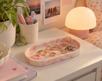 May include: An oval-shaped tray with a pink and white marbled pattern holds a gold chain necklace, two gold rings, a pink beaded ring, a translucent pink hair claw clip, and a small hair clip with a black and white spotted pattern.