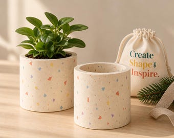 May include: Two cylindrical planters with a cream-colored base and multicolored terrazzo-style flecks. One planter holds a green leafy plant, while a beige drawstring pouch with the text "Create Shape Inspire." sits beside them.
