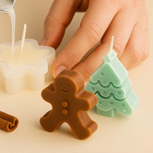 Könnte beinhalten: Eine Person gießt Flüssigkeit in eine sternförmige Form mit einem Kerzendocht. Das Bild zeigt auch eine braune Lebkuchenmann-Kerze, eine grüne Weihnachtsbaum-Kerze und einen Zimtstange. Die Kerzen befinden sich auf einer hellbeigen Oberfläche.