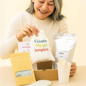 Op de afbeelding: Een doe-het-zelf knutselpakket met een wit tasje met trekkoord en de tekst Create Shape Inspire, een zilverkleurig zakje met het label RESIN, een bruin papieren zakje met een venster met kleurrijke fragmenten en een papieren beker.