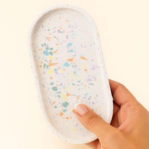 Peut inclure: Un plateau décoratif ovale avec un motif moucheté. Le plateau est blanc avec des éclats colorés de bleu, orange, jaune et violet. Le plateau est tenu par la main d'une personne.