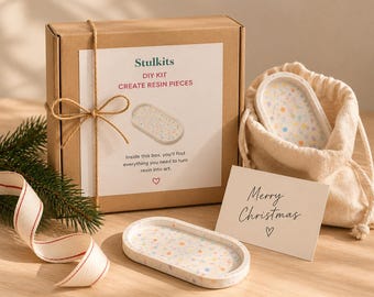May include: A Stulkits DIY resin kit in a brown cardboard box tied with twine, featuring a white label with text. Includes an oval tray with a multicolored confetti pattern, a matching tray in a beige drawstring bag, and a Merry Christmas card.