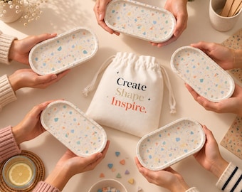 May include: Five oval trays with a multicolored terrazzo pattern featuring flecks of blue, orange, and yellow. A central off-white drawstring fabric pouch displays the text Create Shape Inspire. Small colorful geometric shapes are scattered nearby.