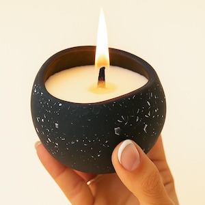 May include: A lit black candle in a speckled, round container. The candle has a white wax interior and a wooden wick. The flame is tall and bright. The container is held in a hand with a French manicure.
