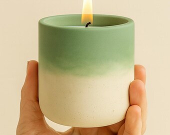 Peut inclure: Une bougie cylindrique dans un récipient en céramique bicolore présentant une partie supérieure vert sauge mat qui se fond dans une base crème mouchetée. La bougie possède une mèche allumée avec une flamme jaune visible et lumineuse.