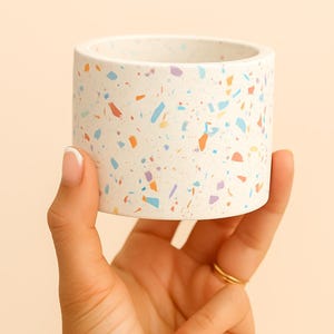Könnte beinhalten: Ein zylindrischer Pflanzentopf mit einem gesprenkelten Terrazzo-Design. Der cremefarbene Keramiktopf hat bunte Flecken in Orange, Blau, Lila und Gelb. Der kleine, runde Topf wird in einer Hand gehalten.