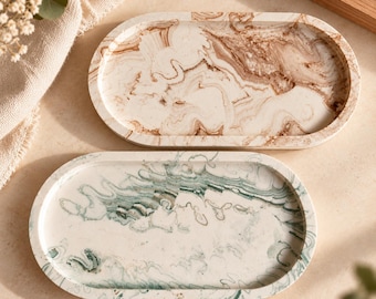 May include: Two oval-shaped trays with raised edges. The top tray features a cream base with swirling brown marble-like patterns. The bottom tray features a cream base with swirling green marble-like patterns. Both trays have a smooth finish.