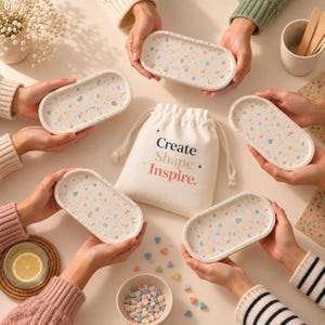 May include: Five oval trays with a multicoloured terrazzo pattern featuring flecks of blue, orange, and yellow. A central off-white drawstring fabric bag displays the text "Create Shape Inspire." Small colourful geometric shapes are scattered nearby.