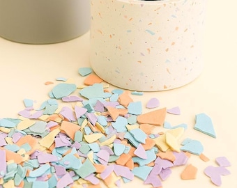 May include: Two cylindrical planters, one gray and one white with colorful flecks, sit on a light surface. Scattered around the planters are pastel-colored, irregularly shaped pieces. The planters are likely for indoor plants.