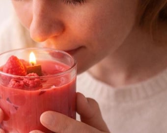 May include: A clear glass candle container filled with pink wax and topped with several whole red raspberries. A single lit wick burns in the center of the wax, emitting a warm glow. The candle is held in a pair of hands, showcasing the vibrant fruit-like texture.