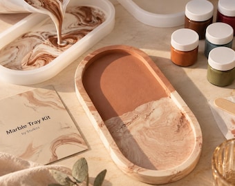 May include: A Marble Tray Kit by Stulkits featuring an oval tray with a two-tone finish of solid terracotta and a marbled beige pattern. The kit includes a white mixing pitcher pouring liquid into a mold, small paint jars, and a printed instruction card.