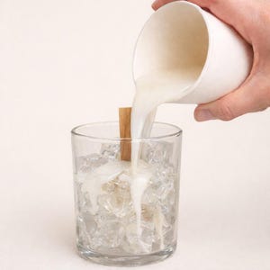 Può includere: Un bicchiere trasparente pieno di cubetti di ghiaccio e un bastoncino di cannella. Un liquido bianco viene versato da una tazza bianca nel bicchiere. Lo sfondo è bianco.