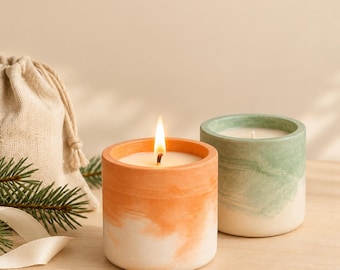 May include: Two cylindrical candles in concrete vessels. One candle is lit, featuring a white and orange marbled finish. The second candle is unlit, with a white and sage green marbled finish. A small beige fabric drawstring pouch and a sprig of pine needles are visible.