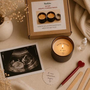 May include: A candle kit with the text "CREATE 2 CANDLE PIECES" and a lit candle that says "WE'RE HAVING BABY". An ultrasound image and a pen with a heart-shaped top are also visible. The kit includes a box with the text "Inside this box, you'll find everything you need to turn wax into art."