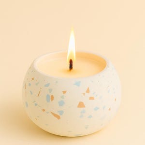 Può includere: Una candela accesa in un contenitore rotondo bianco maculato. La candela ha una cera beige e una lunga fiamma gialla. Il contenitore ha accenti in stile terrazzo blu e arancione. Lo sfondo è giallo chiaro uniforme.
