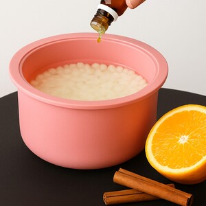 May include: A pink bowl filled with small, white beads, with essential oil being poured from a brown bottle. An orange slice and cinnamon sticks are on a black surface, suggesting aromatherapy or spa products.