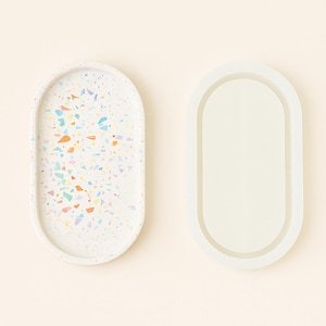 Pode incluir: Duas bandejas ovais. Uma é branca com manchas coloridas em estilo terrazzo. A outra é branca lisa, com uma borda elevada. Ambas estão sobre um fundo creme. Estas bandejas são adequadas para decoração de casa.