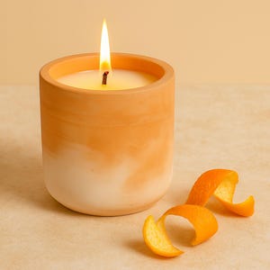 Può includere: Una candela arancione accesa in un portacandele color terracotta con base bianca. La candela ha una fiamma luminosa ed è accanto a riccioli di scorza d'arancia. La candela è su una superficie beige.
