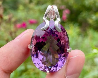 May include: A large, faceted amethyst pendant with a silver bail. The oval-shaped gemstone displays shades of purple and violet. The pendant is held up against a blurred green and pink background.