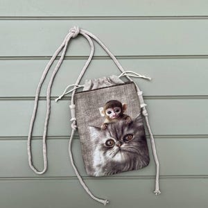 May include: A small, drawstring bag with a light beige rope strap. The bag features a grayscale image of a monkey perched on a cat's head. The bag is made of a textured, linen-like material.