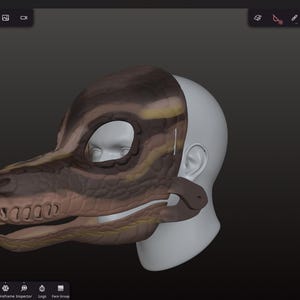 Op de afbeelding: Een 3D-geproduceerd dinosaurusmasker in bruin-, beige- en geeltinten, dat gedeeltelijk een witte mannequinhoofd bedekt. Het masker heeft een gedetailleerd reptielachtig ontwerp met oog- en mondopeningen en een riem.