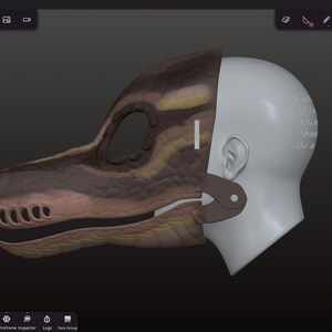 Op de afbeelding: Een 3D-model van een dinosaurus hoofdmasker, met een gedetailleerd bruin en geel getextureerd exterieur. Het masker is gedeeltelijk bevestigd aan een wit, gelaatloos menselijk hoofdmodel, dat het ontwerpproces laat zien. De achtergrond is donkergrijs.