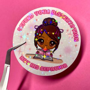 May include: A round, holographic sticker with a cartoon illustration of a person reading a book. The sticker has the text "DEFEND YOUR DISSERTATION NOT HIS BEHAVIOR" in pink. A pair of tweezers is holding the sticker against a pink background.