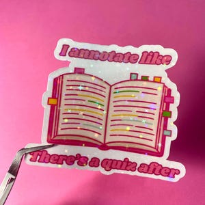 May include: A sticker featuring an open book with annotations and the text "I annotate like" and "There's a quiz after." The sticker has a pink and white border and is held by tweezers against a pink background. The book has colorful tabs.