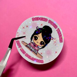 May include: A round, holographic sticker with a cartoon girl reading a book. The sticker has the text "DEFEND YOUR THESIS" and "NOT HIS BEHAVIOR" in pink. The background is pink.