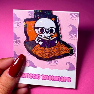 May include: A magnetic bookmark featuring a cartoon skeleton reading a book, wearing glasses, and resting on an orange pillow with a spiderweb design. The bookmark has a holographic finish and the text "Magnetic Bookmark" on a pink background.
