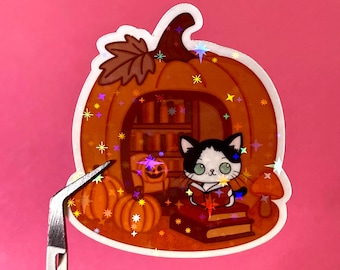 May include: A holographic sticker featuring a black and white cat reading a book inside a pumpkin-shaped library. The sticker has a fall theme with pumpkins, a mushroom, and a bookshelf. The sticker is on a pink background.