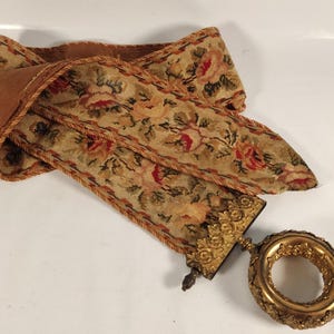 May include: An antique tapestry ribbon with a floral pattern in red, pink, and green on a beige background. The ribbon is trimmed with a gold-coloured rope and attached to a decorative gold-coloured ring.