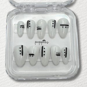 May include: A set of ten white oval-shaped press-on nails with black minimalist designs. Each nail features a combination of vertical lines and small dots. The nails are displayed in a clear plastic case, with the brand name "poppies" printed on the card.
