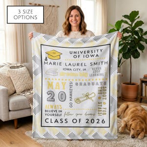 May include: A white graduation blanket with a black and yellow geometric border. The blanket features text including "University of Iowa," "Marie Laurel Smith," "Class of 2026," and "May 20." The blanket is held by a person.