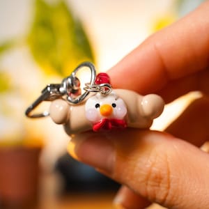 Può includere: Un portachiavi fatto a mano con una figura di gallina in miniatura. La gallina ha la testa bianca, un bargiglio rosso e un becco arancione. Il portachiavi è realizzato in pasta polimerica e ha una chiusura color argento.