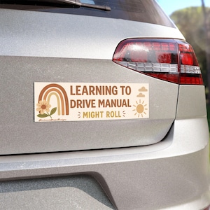 Op de afbeelding: Een rechthoekige bumpersticker met een crèmekleurige achtergrond en de bruine tekst LEARNING TO DRIVE MANUAL MIGHT ROLL. Het ontwerp bevat een bruine regenboog, een bloem, twee wolken en een zonicoon. De sticker is aangebracht op een zilveren auto.
