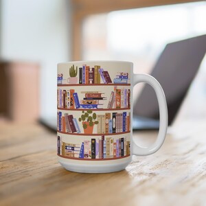 Puede incluir: Taza de cerámica blanca con una ilustración de una estantería llena de libros coloridos. El diseño incluye plantas en macetas y la palabra "Tofein" en el lomo de un libro. La taza tiene un asa grande y está sobre una superficie de madera.
