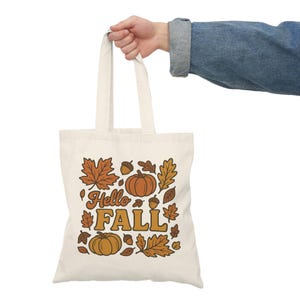 May include: Cream-colored tote bag with a fall-themed design. The bag features the words "Hello FALL" in orange and brown, surrounded by illustrations of pumpkins, leaves, and acorns. The bag is held by a person wearing a denim jacket.