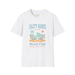 Puede incluir: Camiseta blanca con un diseño de playa. El diseño incluye el texto "SALTY BABES Est. 2025" sobre una palmera, olas, sol y dos tumbonas. Debajo, se lee "Beach Club" y "Girls just wanna have sun."