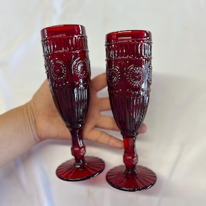 May include: Two deep red glass champagne flutes featuring a textured pattern of vertical ribs and circular floral motifs. Each glass has a slender stem with a decorative knob and a wide, ribbed circular base, crafted from translucent ruby glass.