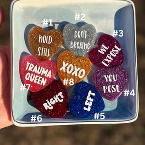 May include: A collection of eight glittery heart-shaped pins with various phrases in white text, including "Hold Still," "Don't Breathe," and "We Expose." The pins are arranged in a square, light blue dish.