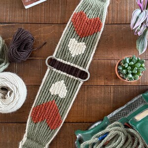 May include: A woven horse cinch in shades of green, cream, and rust, featuring heart and leaf patterns. The cinch has metal hardware and is displayed on a wooden surface with books and other equestrian-themed items.