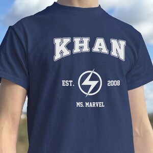May include: Navy blue t-shirt featuring the white text "KHAN" above a white lightning bolt symbol. Below the symbol are the words "EST. 2008" and "MS. MARVEL". The shirt has short sleeves.