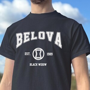 May include: Dark navy blue t-shirt with the text "BELOVA" in white arched lettering above a circular graphic. Below the graphic are the words "EST. 1989 BLACK WIDOW" in white. The shirt is short-sleeved.