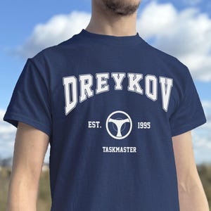 May include: Navy blue t-shirt with white text that reads "Dreykov" above a steering wheel graphic. Below the graphic are the words "EST. 1995" and "TASKMASTER". The shirt is short-sleeved.